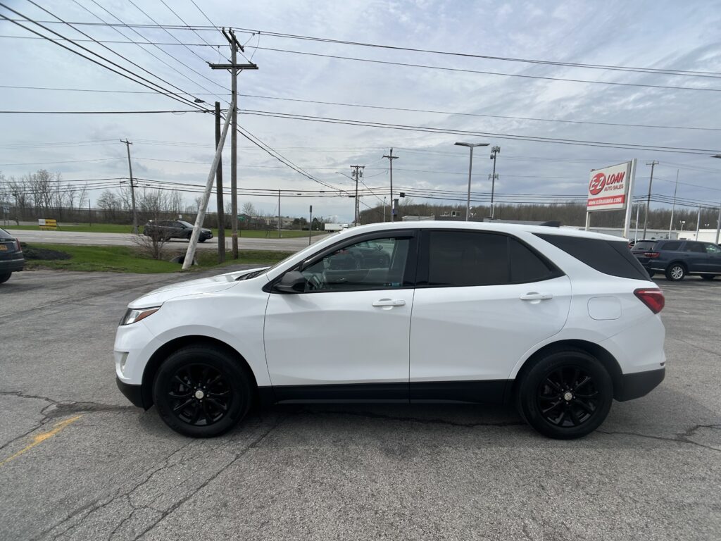 Side view of a white crossover SUV with black wheels parked in a dealership lot on a cloudy day, with an EZ Loan Auto Sales sign in the background.