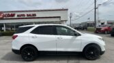 Side view of a white SUV with black wheels parked in front of a car dealership with a red LOAN AUTO SALES sign in the background.