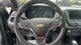 Front interior view of a Chevrolet showing the steering wheel, instrument cluster, and center touchscreen display. tactile details include the Chevy logo on the wheel and visible controls on the spokes, with the gauge cluster behind it and the gear selector nearby in the console.