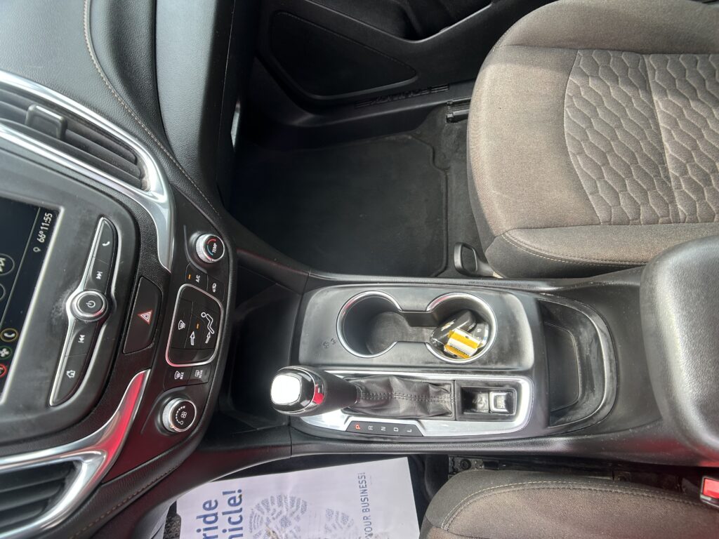 Interior of a car front cabin: center console with gear selector, cup holders, and dashboard controls visible behind the driver seat rail.