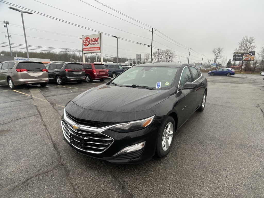 Black Chevrolet sedan parked in a rainy dealership lot with other minivans nearby and an EZ Loan Auto Sales sign in the background.