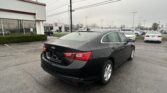 Rear three-quarter view of a black Chevrolet sedan parked in a car dealership lot on a cloudy, rainy day with other cars and a showroom in the background.