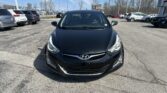 Front view of a black Hyundai in the foreground of a car dealership lot, with several SUVs and a bright blue sky in the background.