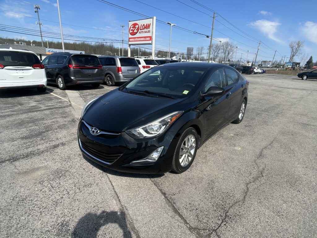 Black Hyundai sedan in the foreground at a car dealership lot with row of minivans behind and an E-Z Loan Auto Sales sign against a blue sky