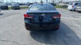 Rear-view of a black Hyundai Elantra parked in a dealership lot with other vehicles and a Toyota sign in the background.