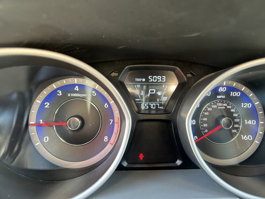 Driver's view of the instrument cluster: left tachometer, right speedometer, center digital display shows Trip A 509.3 and odometer 65,707 miles with a red warning light on bottom.