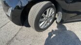 Close-up of a black car's front wheel and fender, showing a silver alloy wheel and treaded tire with a headlight at the left edge, parked on asphalt.