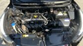 Front view of car engine bay with hood open, showing the battery on the right and various hoses and components in the engine compartment.