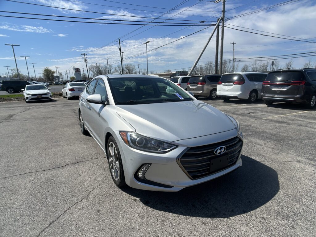 Silver Hyundai sedan in the foreground at a car dealership lot, with rows of vans and SUVs under a clear blue sky and power lines overhead.