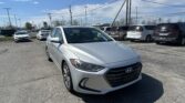 Silver Hyundai sedan in the foreground at a car dealership lot, with rows of vans and SUVs under a clear blue sky and power lines overhead.