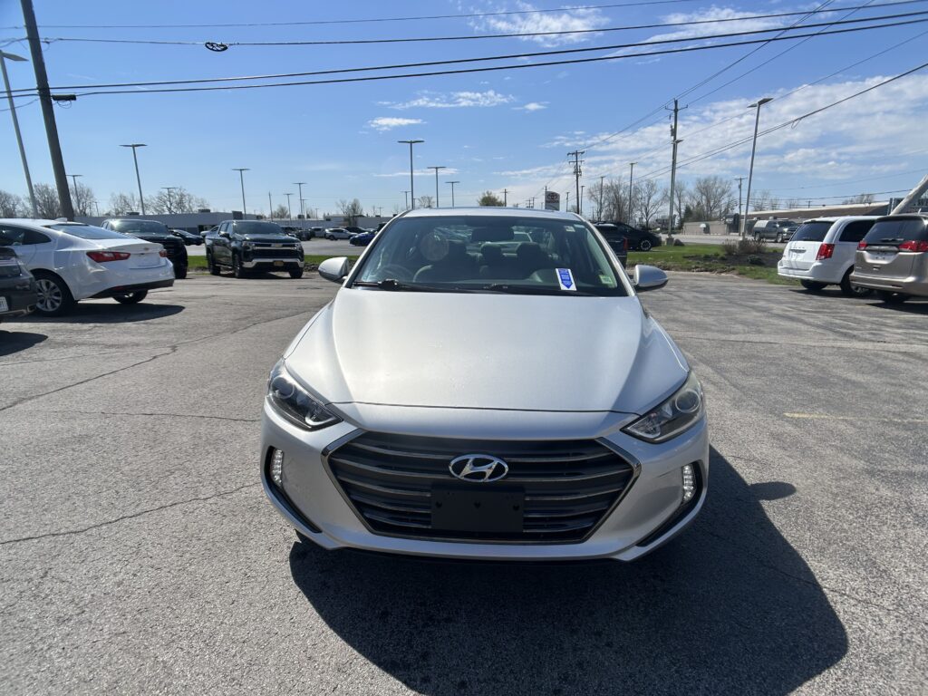 Front view of a silver Hyundai in a car dealership lot with other cars and power lines overhead on a sunny day.