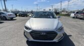 Front view of a silver Hyundai in a car dealership lot with other cars and power lines overhead on a sunny day.