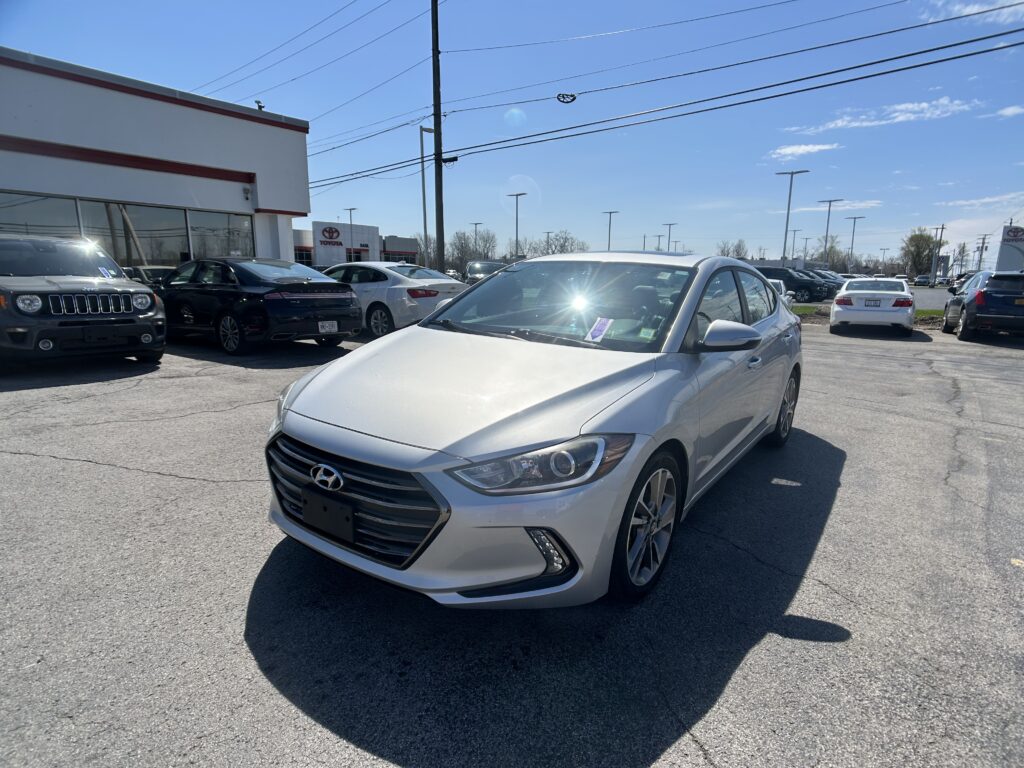 Silver Hyundai sedan in the foreground at a car dealership lot, with a white building on the left and other cars behind it.