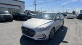 Silver Hyundai sedan in the foreground at a car dealership lot, with a white building on the left and other cars behind it.