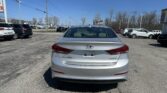 Rear view of a silver Hyundai Elantra Limited in a car dealership lot on a sunny day with other vehicles and a blue sky behind.