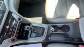 Front passenger area of a car showing the gear selector, climate and media controls, and a beige perforated seat with papers on the floor/seat nearby.