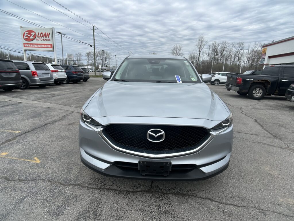 Front view of a silver Mazda SUV parked in a dealership lot, with an E-Z LOAN AUTO SALES sign visible on the left and rows of other vehicles around.