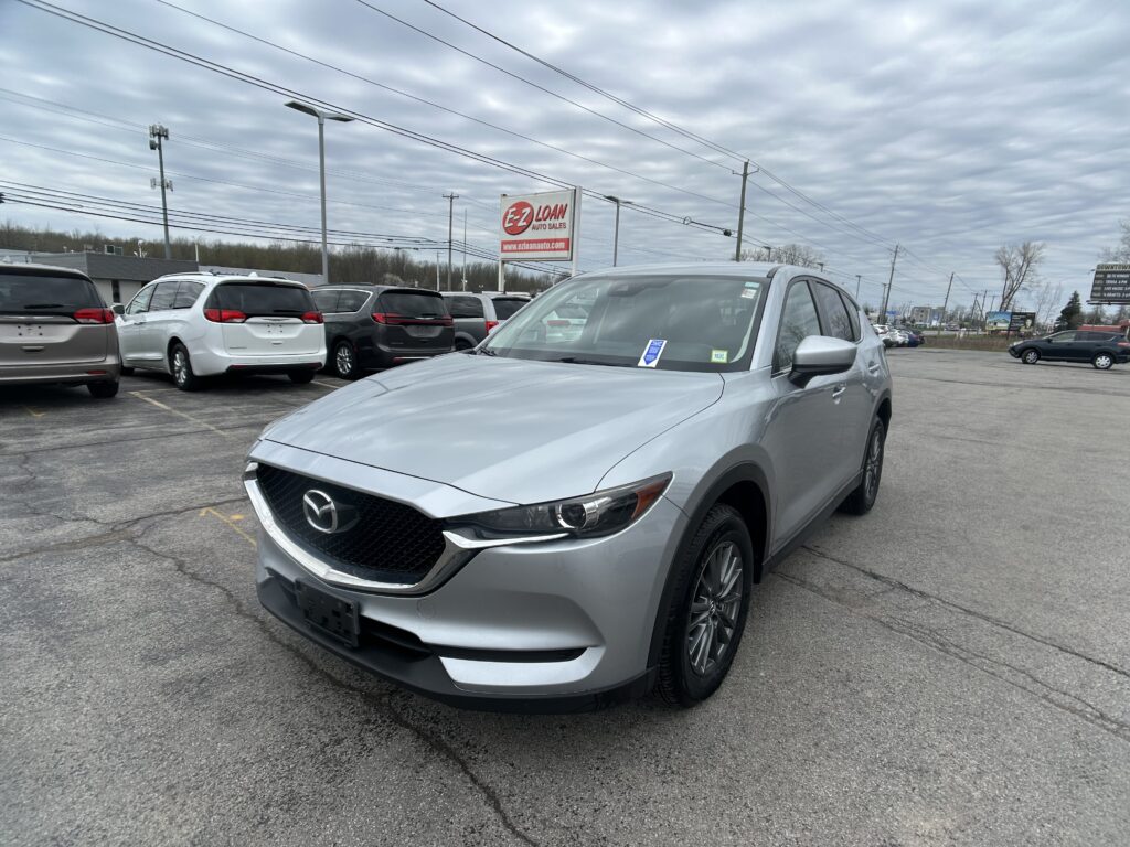 Silver Mazda SUV in a dealership lot with other minivans and an E-Z LOAN sign in the background under an overcast sky.