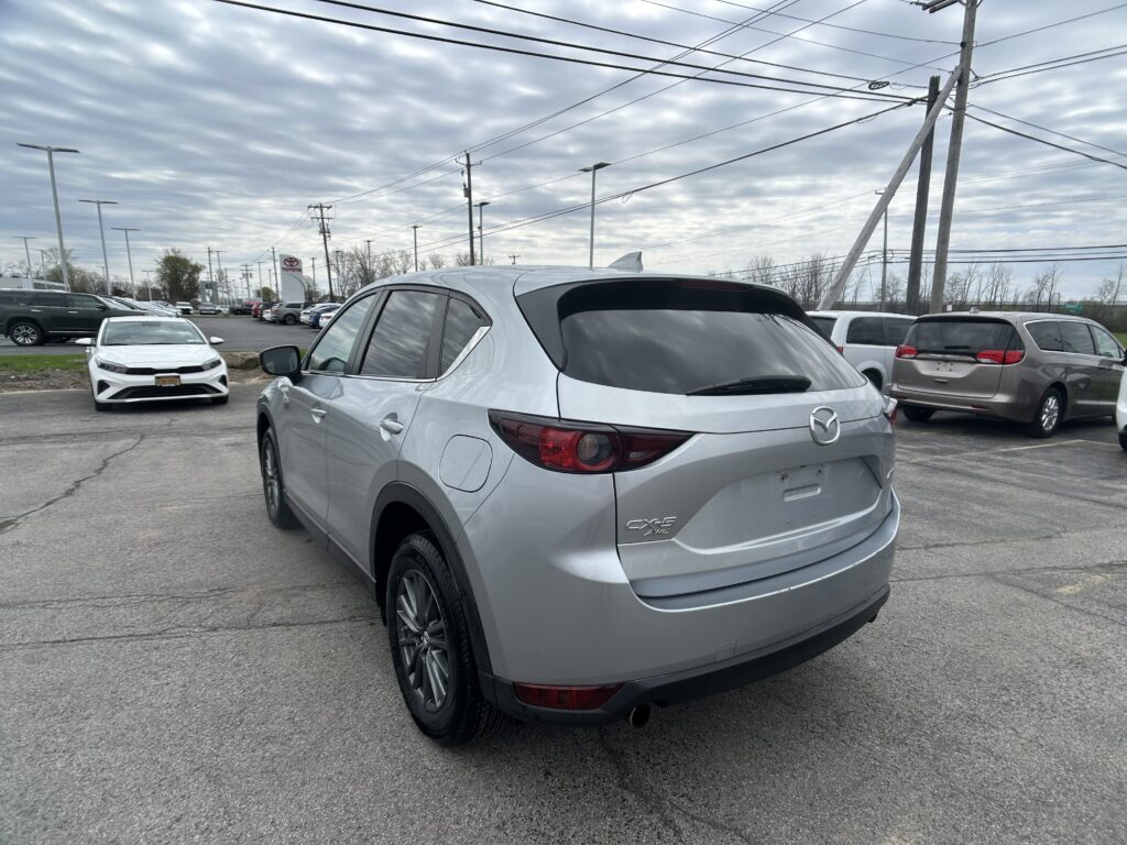 Silver Mazda CX-5 SUV in the foreground in a busy car lot with other vehicles and tall power lines under a cloudy sky.