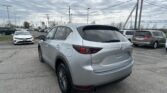 Silver Mazda CX-5 SUV in the foreground in a busy car lot with other vehicles and tall power lines under a cloudy sky.