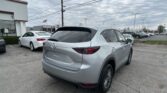 Rear three-quarter view of a silver Mazda SUV in a dealership lot, with a white building to the left and other cars parked in the background under a cloudy sky.