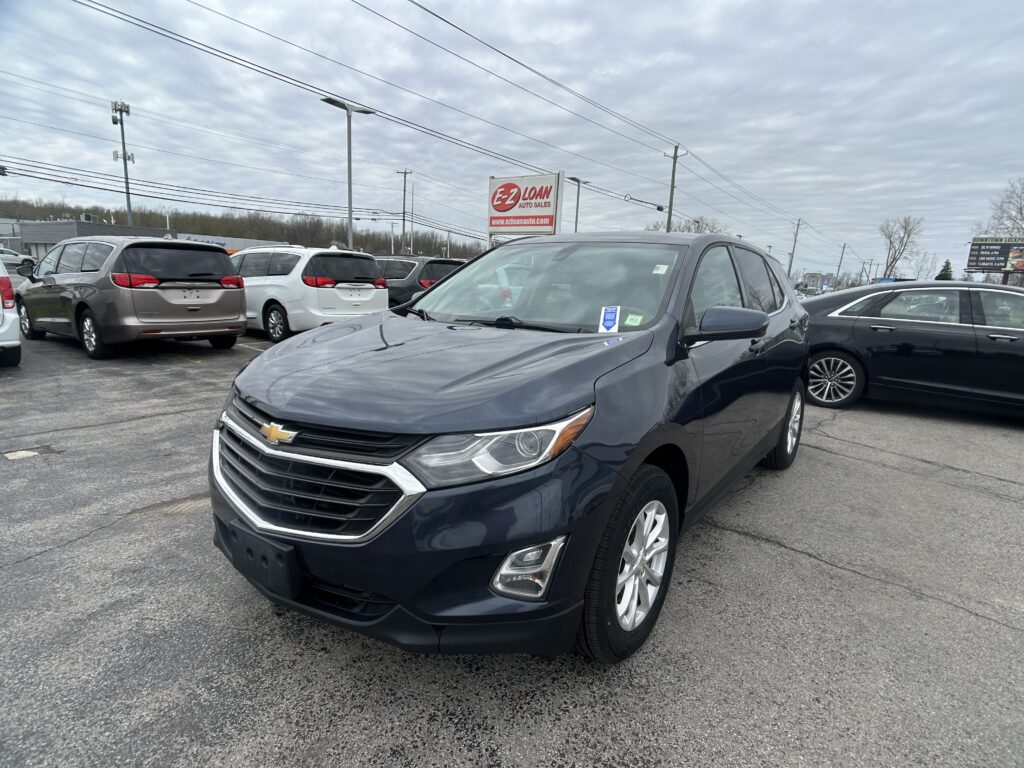 Gray Chevrolet SUV in the foreground at a car dealership lot with multiple vans and a red EZ Loan sign in the background.