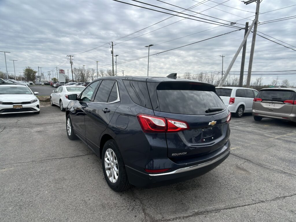 Rear three-quarter view of a dark blue Chevrolet Equinox SUV in a car dealership lot, with other cars and a cloudy sky above.