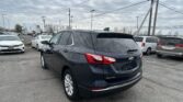 Rear three-quarter view of a dark blue Chevrolet Equinox SUV in a car dealership lot, with other cars and a cloudy sky above.