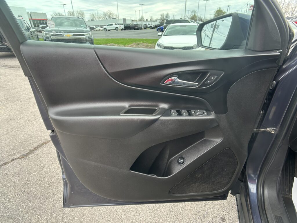 Inside of a car door panel with power window switches and a chrome door handle; view of a dealership parking lot outside.