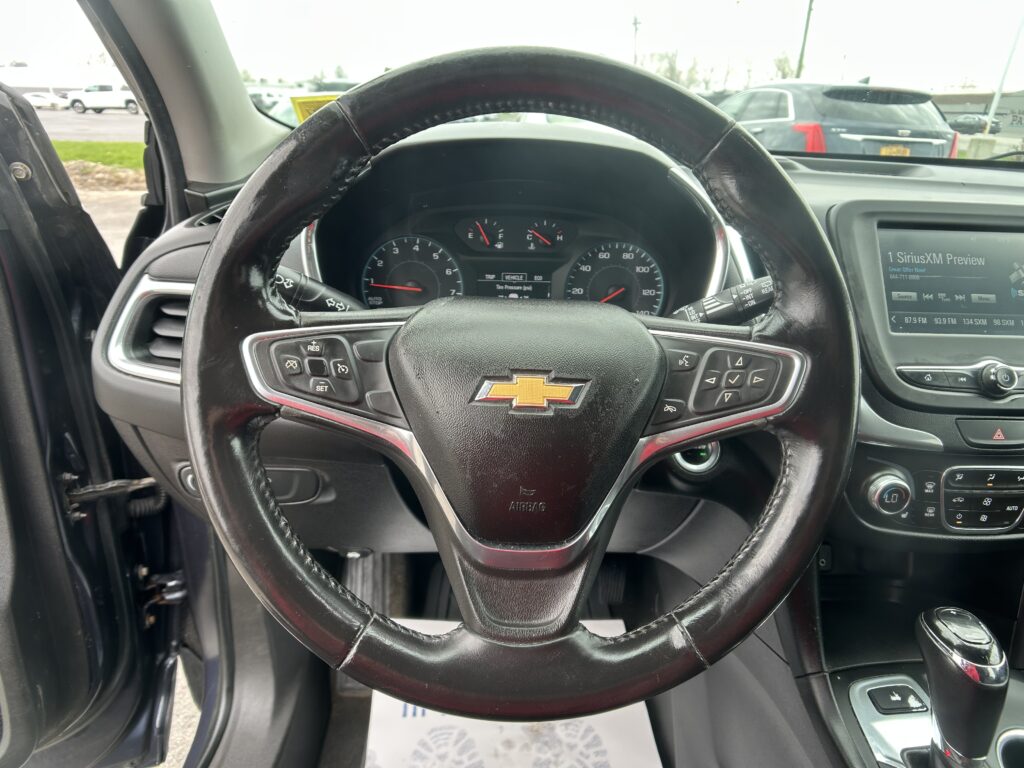 Close-up of a black leather steering wheel with Chevrolet emblem, dashboard gauges behind, and an infotainment screen to the right in a parked car interior