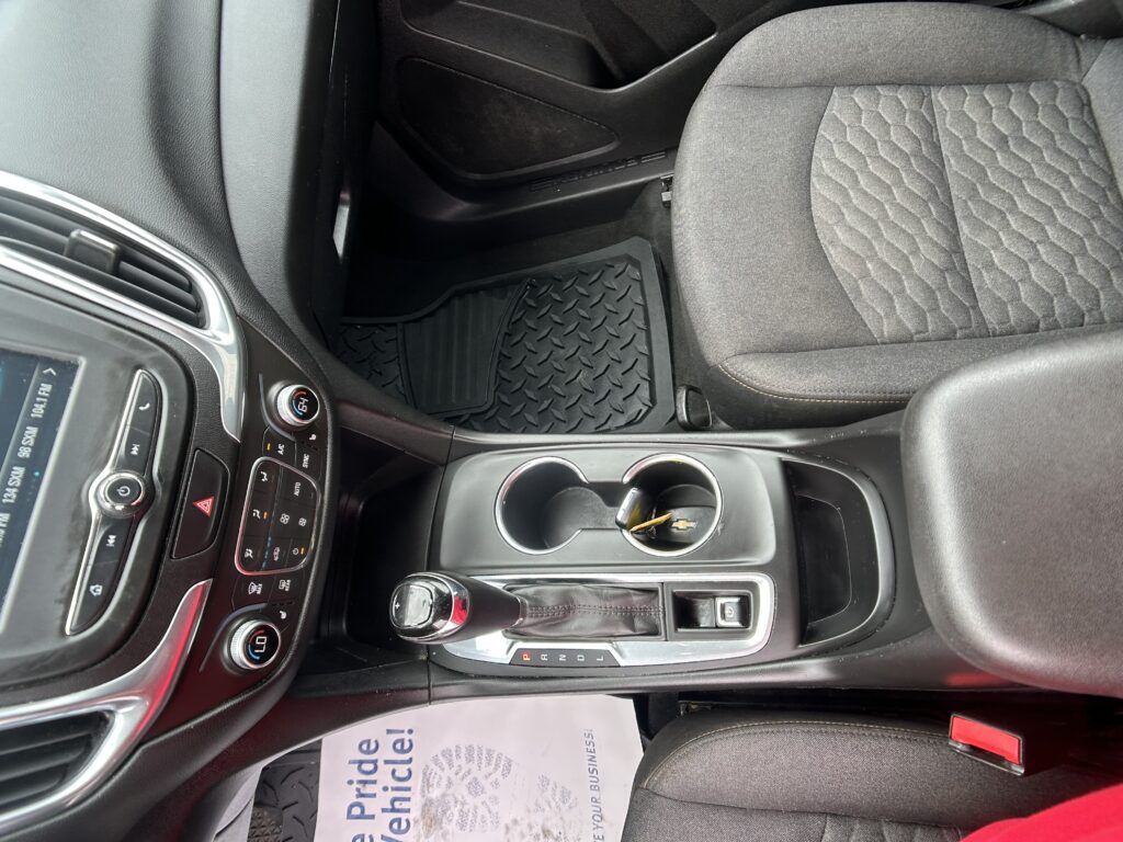 Interior of a car showing the center console, automatic gear shifter, cup holders, and textured seat upholstery with a rubber floor mat nearby