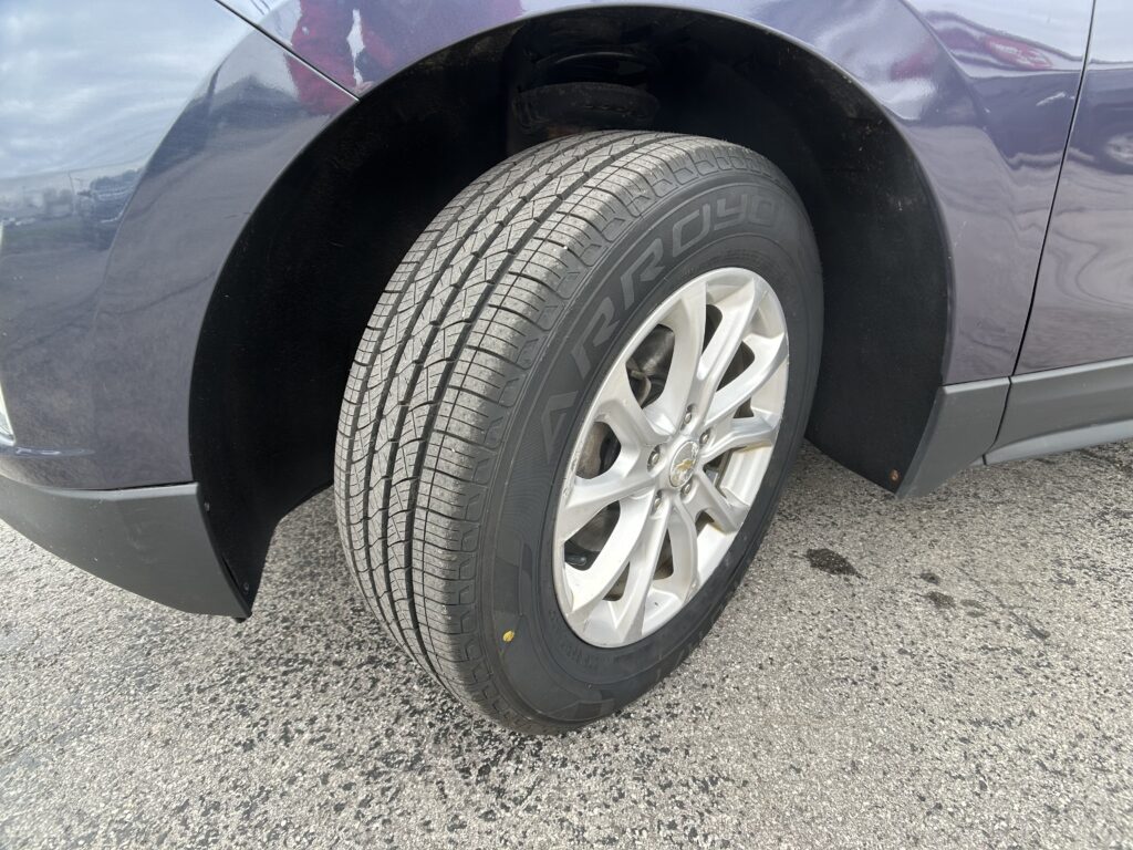 Close-up of a car tire mounted on a silver alloy wheel, with the wheel well and dark blue body nearby on asphalt road it’s parked on.