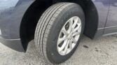 Close-up of a car tire mounted on a silver alloy wheel, with the wheel well and dark blue body nearby on asphalt road it’s parked on.