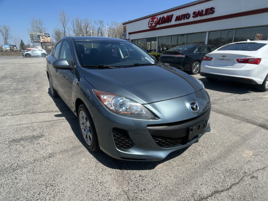 Blue-gray hatchback/compact car in the foreground at an auto dealership lot with other cars and a storefront named E-Z LOAN AUTO SALES in the background.