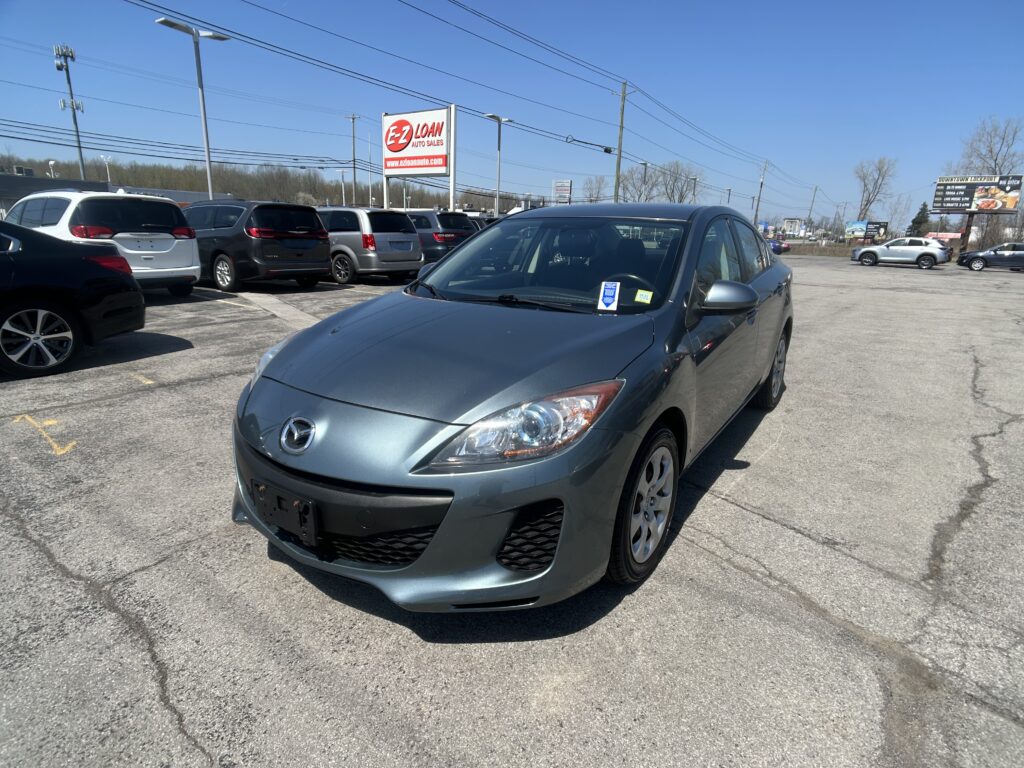 Foreground shows a gray Mazda sedan in a car dealership lot, with multiple minivans and SUVs nearby and an EZ Loan Auto Sales sign in the background on a clear day.