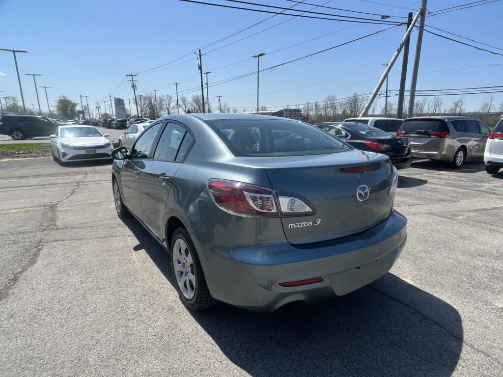 Rear three-quarter view of a silver Mazda3 parked in a busy car lot under a clear blue sky, with other cars and power lines in view.