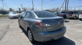 Rear three-quarter view of a silver Mazda3 parked in a busy car lot under a clear blue sky, with other cars and power lines in view.