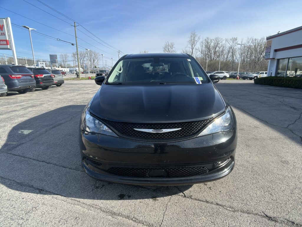 Front view of a dark gray Chrysler minivan parked in a dealership lot on a sunny day, with other cars and a building in the background.