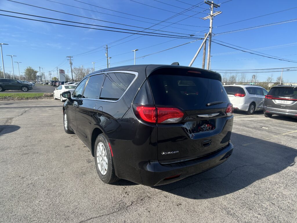 Black Kia Voyager minivan in the foreground at a car dealership lot with other vehicles and power lines overhead, clear blue sky above.
