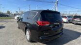 Black Kia Voyager minivan in the foreground at a car dealership lot with other vehicles and power lines overhead, clear blue sky above.