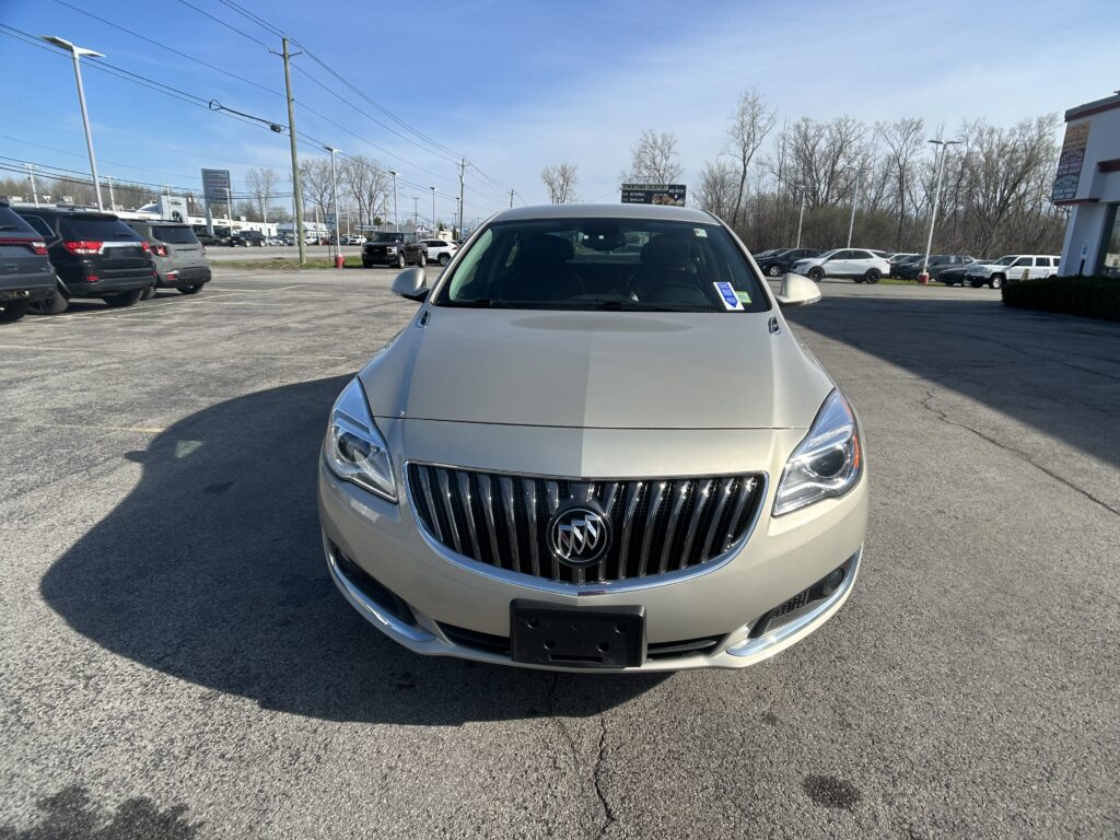 Beige Buick SUV seen from the front in a dealership lot with other cars and leafless trees in the background.”