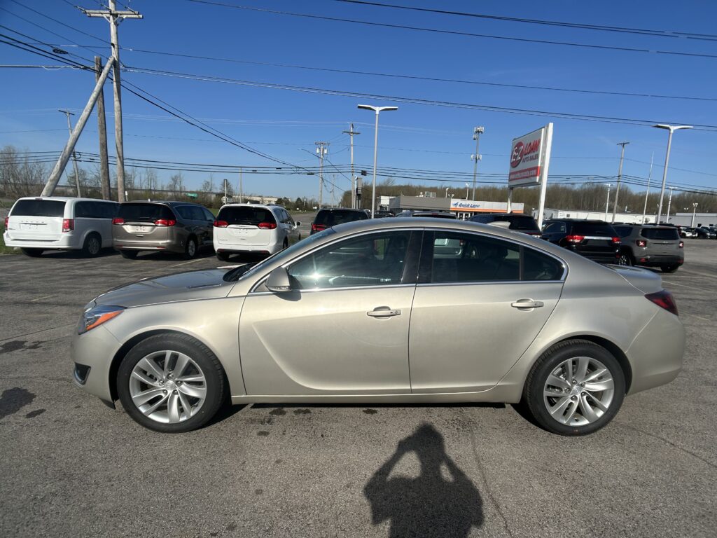 Front corner of a silver sedan in a car dealership lot with other minivans parked in the background and power lines overhead under a clear blue sky.