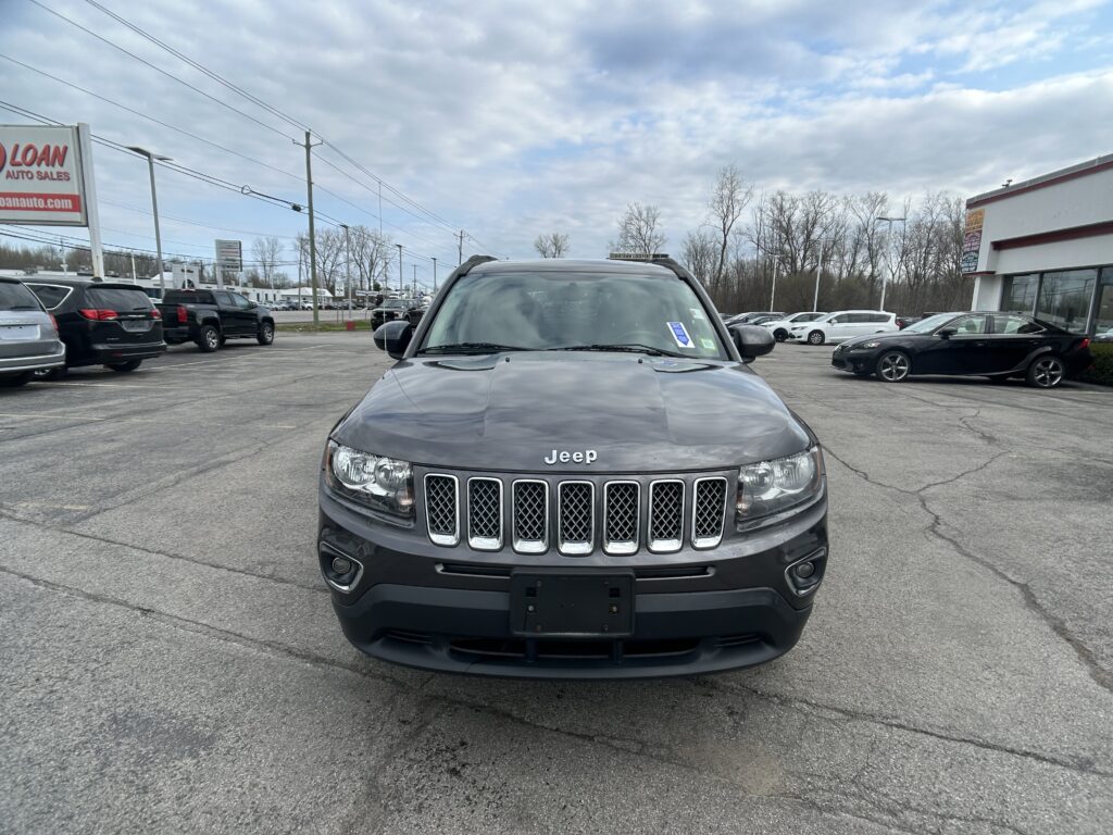 Front view of a gray Jeep SUV in a dealership lot with other cars and a car-sales sign in the background.