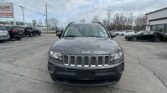 Front view of a gray Jeep SUV in a dealership lot with other cars and a car-sales sign in the background.