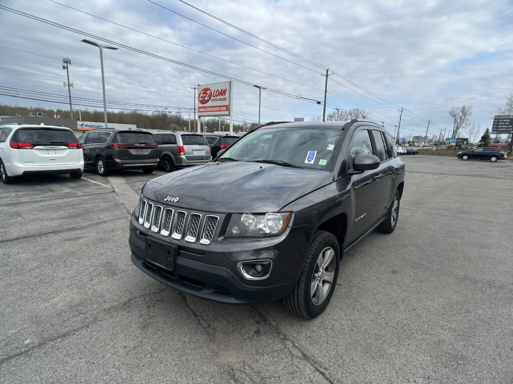 Front-right view of a gray Jeep SUV in a car dealership parking lot, with other vehicles and an EZ Loan Auto Sales sign in the background under a cloudy sky.