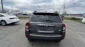 Rear view of a gray Jeep SUV in a parking lot with several cars and a cloudy sky behind it.