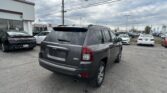 Front and center, a dark gray Jeep SUV in a car dealership lot with other vehicles and a white building to the left on a cloudy day.