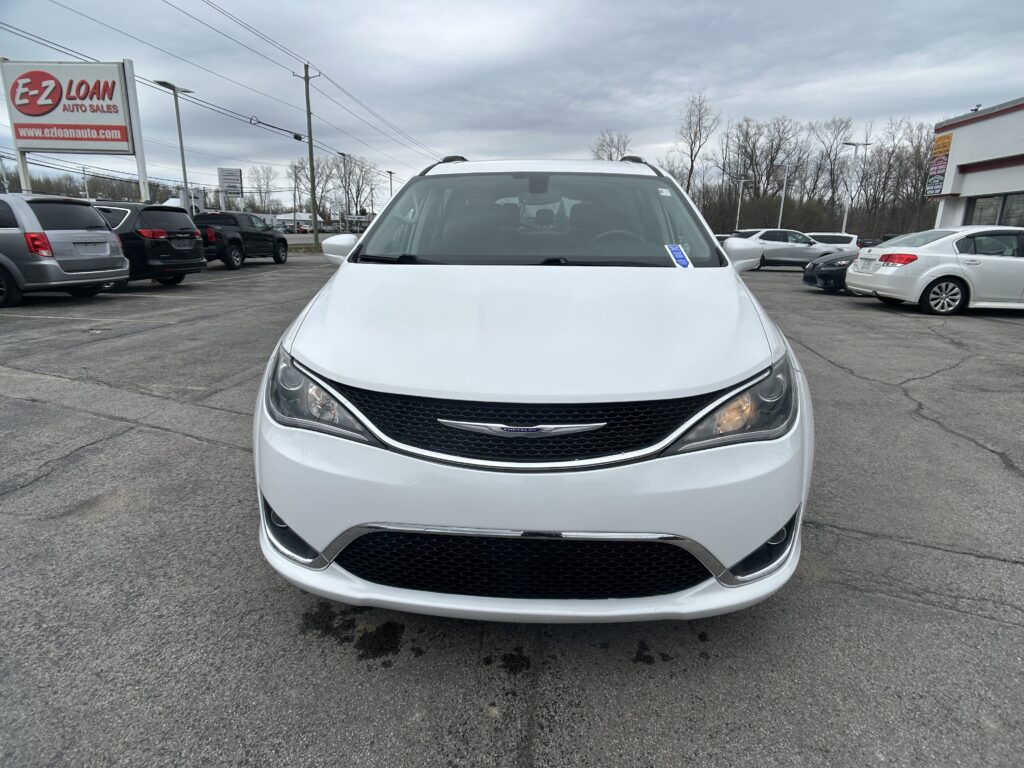 Front view of a white Chrysler minivan parked in a dealership lot with other cars and a large E-Z Loan sign in the background.