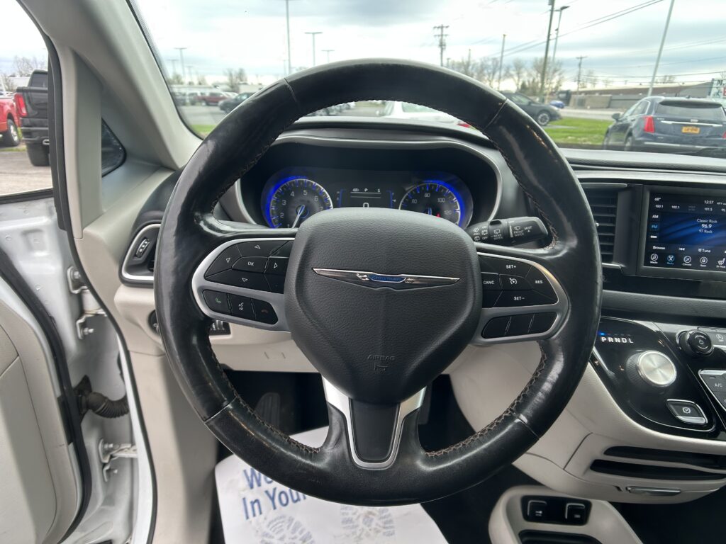 Driver’s view from the driver’s seat showing a black steering wheel, illuminated blue dashboard gauges, and a car interior in a parking lot outside the windshield.