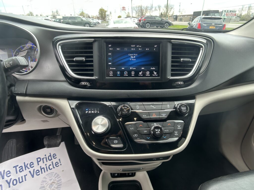 Center car dashboard with touchscreen display showing radio and climate controls; parking lot visible through windshield outside the car.